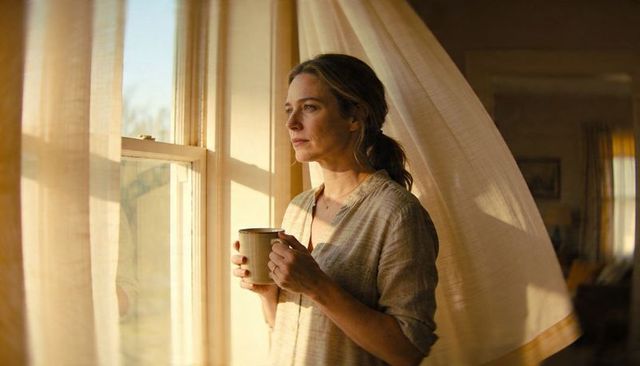 Holding ceramic mug and gazing through sunlit sash window, woman wearing linen blouse