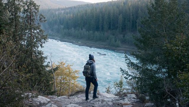 Standing hiker observing turquoise river rapids from rocky forest overlook, backpacking