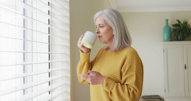 Senior woman in yellow sweater drinking coffee and looking outside through window with shades. Suitable for content on daily routines, relaxation, healthy living, and retirement lifestyle.