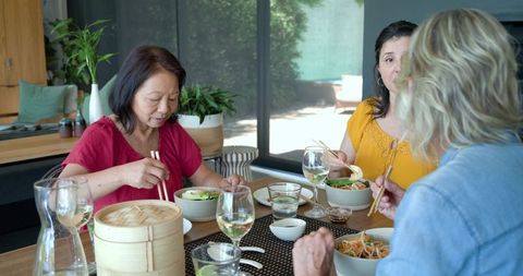 Diverse women sharing asian-style meal with chopsticks at sunlit modern dining table