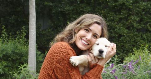 Mid-adult woman cradling golden retriever puppy, smiling in garden with purple flowers