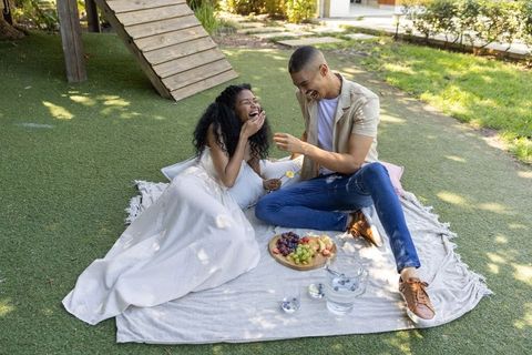 Joyful diverse couple laughing on picnic blanket sharing fruit and daisy in sunlit backyard