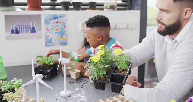 Diverse male teacher and happy schoolchildren studying plants and wind turbines in school classroom. Education, inclusivity, elementary school and learning concept.