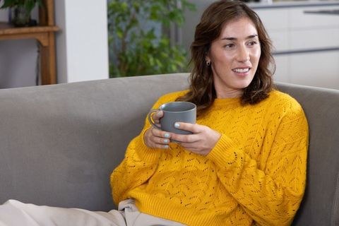 Woman relaxing on sofa holding mug wearing mustard yellow knit sweater smiling