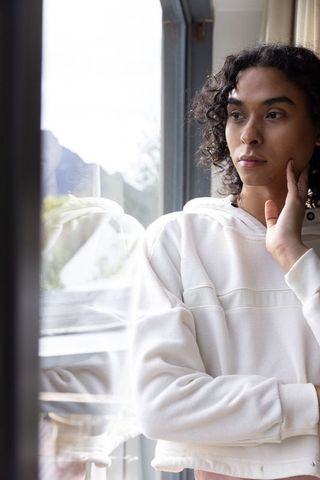 Pensive nonbinary adult gazing through window wearing white hoodie