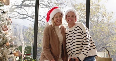 Mother daughter wearing santa hats smiling by window with festive christmas tree