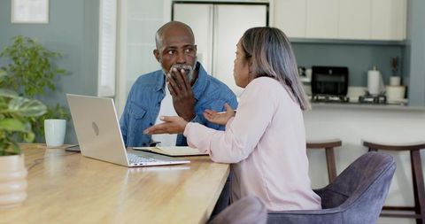 Senior Diverse Couple Discussing Finances at Kitchen Table with Laptop and Notebook