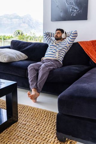 Indian man relaxing on navy sectional sofa wearing headphones in bright living room