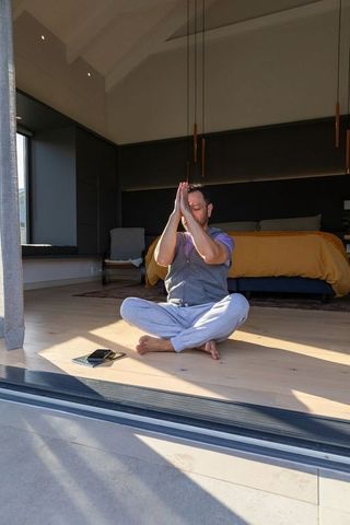 Mature asian man meditating on sunlit modern bedroom floor near open sliding door
