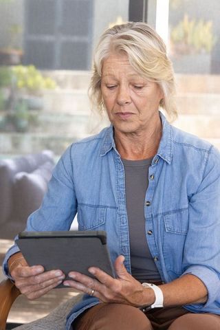 Senior woman using tablet and wearing smartwatch while sitting in sunlit cozy living room