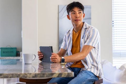 Asian young man sitting at kitchen island holding tablet, looking thoughtful, casual sunlit