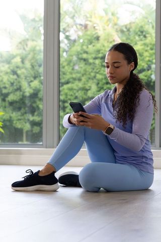 African american woman sitting by sunlit window checking phone in pastel workout gear