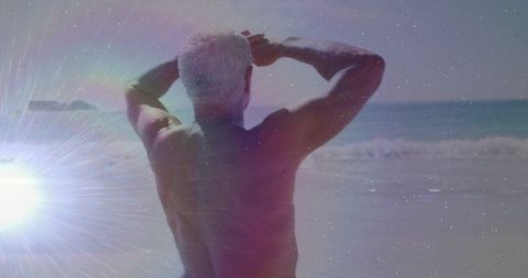 Senior man standing shirtless facing ocean at sunlit beach with lens flare