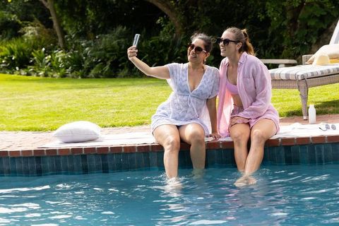 Two friends taking selfie while relaxing at backyard pool, dipping legs, wearing sunglasses