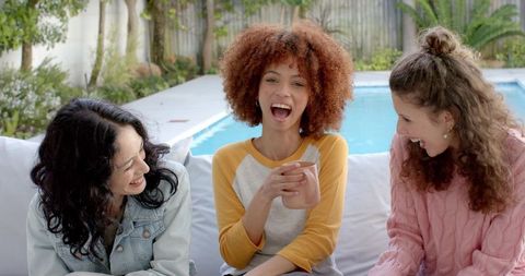 Diverse friends laughing and chatting on outdoor sofa by pool holding mug of coffee