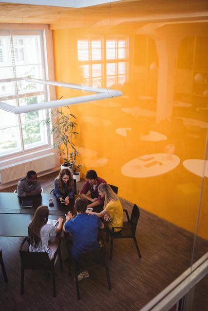 Diverse group of business executives collaborating around a table in a modern office with a bright orange wall. Suitable for illustrating teamwork, professional collaboration, corporate environments, business meetings, and office culture in marketing materials, presentations, and websites.