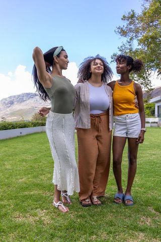 Three diverse women standing arm-in-arm on lawn, smiling and enjoying sunny summer garden