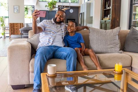 Smiling african american father taking selfie with son on modern living room sofa at home