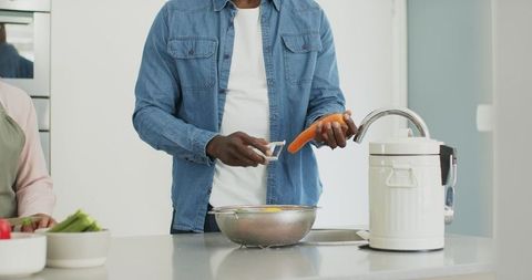 Senior couple home cooking man peeling carrot over colander on modern kitchen island