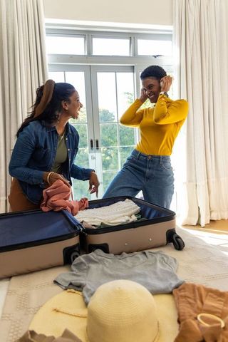 Diverse friends packing suitcase and laughing in sunlit bedroom preparing for trip