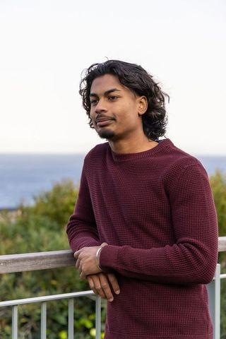 Indian man leaning on railing gazing toward ocean horizon in maroon sweater