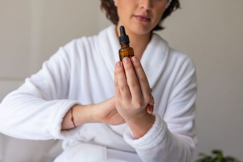 Woman holding amber dropper bottle wearing white robe showing gentle skincare ritual at home