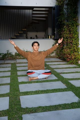 Young asian man sitting cross-legged stretching arms on patio pavers near folding door