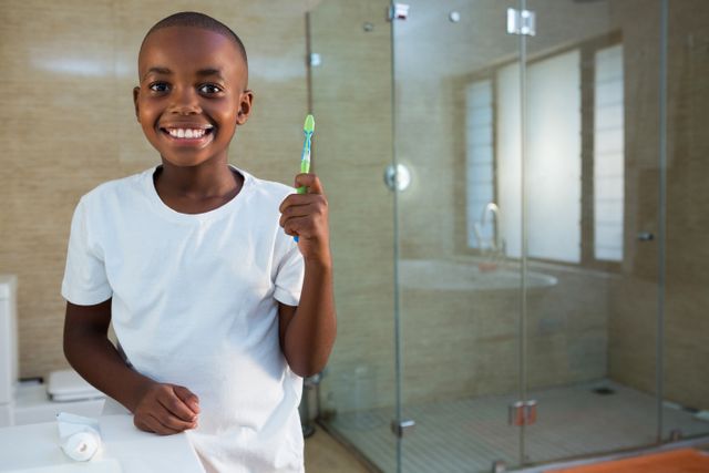 Young boy smiling while holding a toothbrush in a modern bathroom. Ideal for promoting dental hygiene, children's health, morning routines, and personal care products. Suitable for use in educational materials, health campaigns, and advertisements for dental care products.