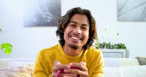 Young man smiling holding mug on cozy mustard sweater couch at home