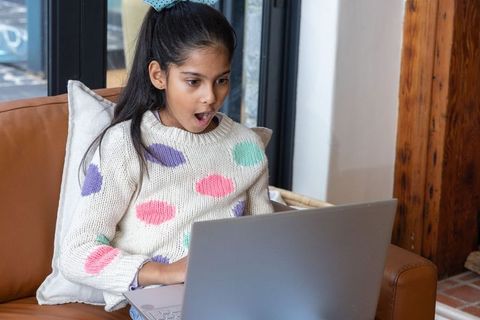 Surprised asian girl reacting to laptop while sitting on couch in cozy sunlit living room