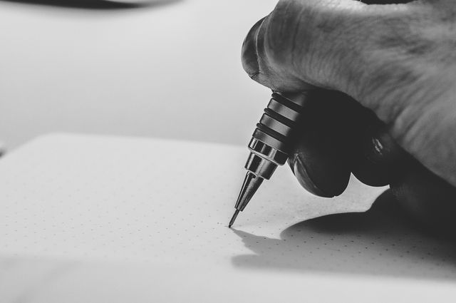 Close-up of a hand holding a pen, writing on dotted paper in black and white. Perfect for illustrating concepts related to handwriting, journaling, note-taking, studying, or office work. Useful for educational materials, productivity blogs, or motivational content about writing.