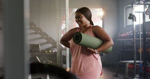 Mid adult african american woman carrying yoga mat in gym in pink athleisure and smiling