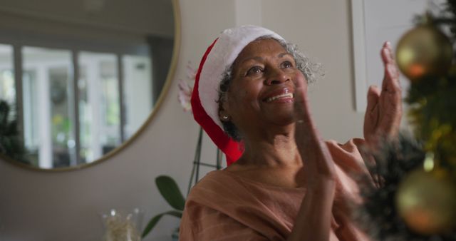 Elderly woman wearing Santa hat smiling at holiday decorations indoors. Ideal for use in holiday greeting cards, senior lifestyle promotions, and festive celebrations.