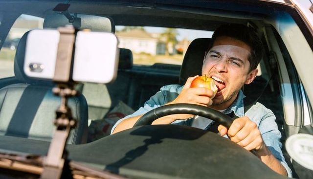 Male driver biting apple and gripping steering wheel with smartphone mounted on dashboard