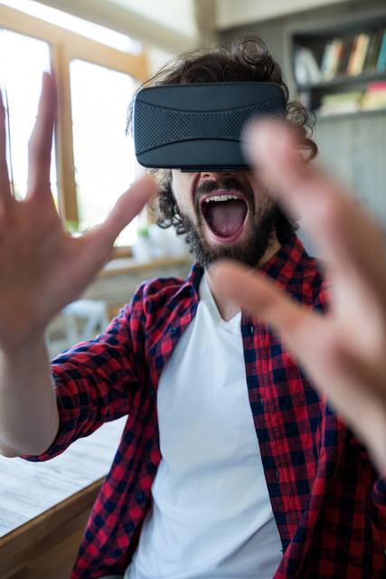 Excited man engaged in a virtual reality experience while sitting in a coffee shop. This image is ideal for showcasing modern technology, digital entertainment, and the immersive nature of VR. It can be used in promotional materials, articles about virtual reality advancements, technology blogs, and advertisements for VR products or coffee shops.