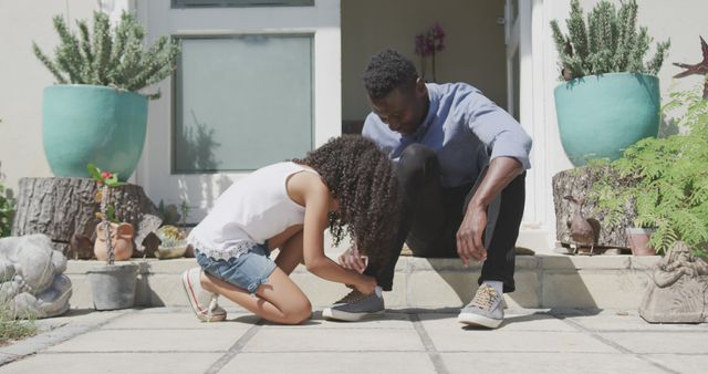 Father tying shoe while daughter helps on house porch. Can be used for family bonding themes, parenting advice articles, outdoor activities depictions, or advertisements promoting family time.