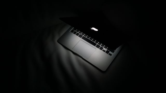 A partially open laptop revealing glowing Apple logo in dark room. Ideal for technology concept, work-from-home setup, night working sessions, computer security, digital illuminated design.