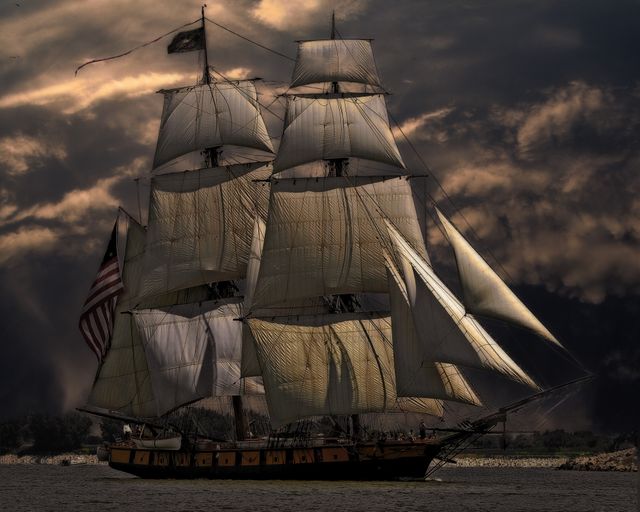 Historical tall ship sailing through the sea under a dramatic, cloudy sky. The image showcases the majestic and meticulously detailed sails, with the ship's dark silhouette contrasting against the sky. Suitable for use in themes related to travel, adventure, nautical, maritime heritage, historical reenactments, and exploration. Perfect for illustrating historical sea voyages, educational content on maritime history, or travel brochures highlighting nautical experiences.