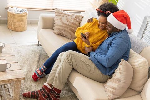 African american couple on couch wearing santa hat and patterned socks, sharing smartphone
