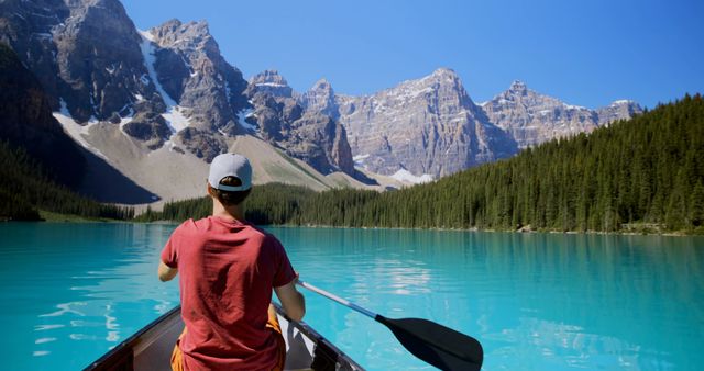 Person paddling a canoe on a clear turquoise lake surrounded by towering snow-capped mountains and dense pine forest. Perfect for promoting outdoor activities, travel destinations, nature retreats, and adventure tourism.