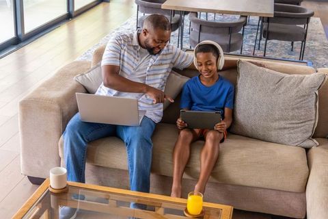 African american father guiding son with tablet on sofa at home, laptop and headphones