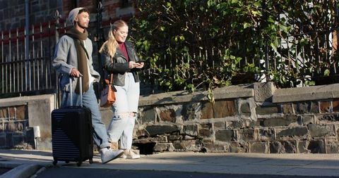 Young couple walking with rolling suitcase on sunlit urban sidewalk checking smartphone