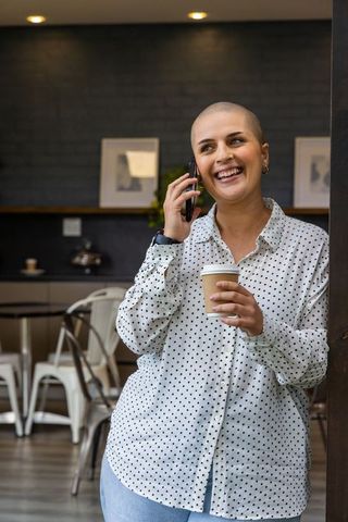 Smiling 30s woman talking on phone holding coffee while leaning on beam at urban cafe