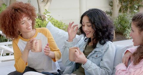 Diverse young women laughing and sharing coffee on outdoor patio sofa