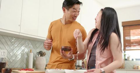 Asian couple laughing and cooking together in bright modern kitchen, sprinkling seasoning