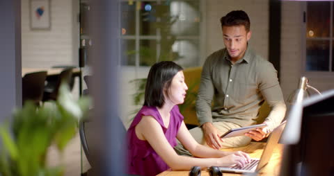 Two coworkers collaborate in a modern office setting, with one viewing a tablet intricately while the other focuses on a laptop. This video captures a dynamic and engaging teamwork process, perfect for illustrating themes of professional collaboration, modern office environments, or technology in business. Useful for business articles, blog posts about workplace productivity, or promotional materials showcasing teamwork.
