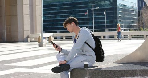 Young man sitting on plaza ledge using smartphone and earbuds, backpack and coffee