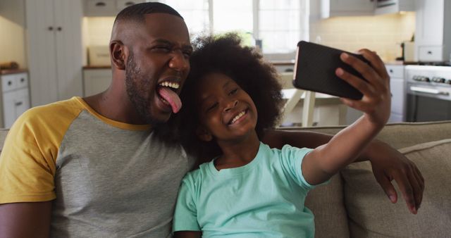 Father and daughter enjoying a fun moment together taking a selfie in their living room. Joyful and loving family scene suitable for use in advertising, family blogs, parenting articles, or social media content promoting bonding and modern technology use in family activities.