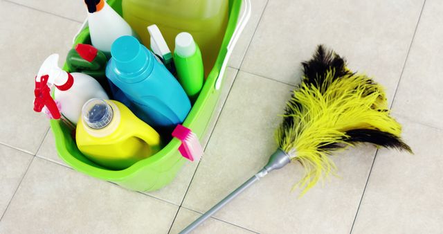 A variety of cleaning supplies, including sprays, detergents, and a feather duster, are gathered in a green bucket on a tiled floor, with copy space. These tools are essential for maintaining cleanliness and hygiene in domestic or commercial environments.