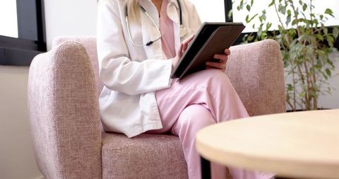 Female physician sitting on pink armchair wearing white coat and pink scrubs holding tablet